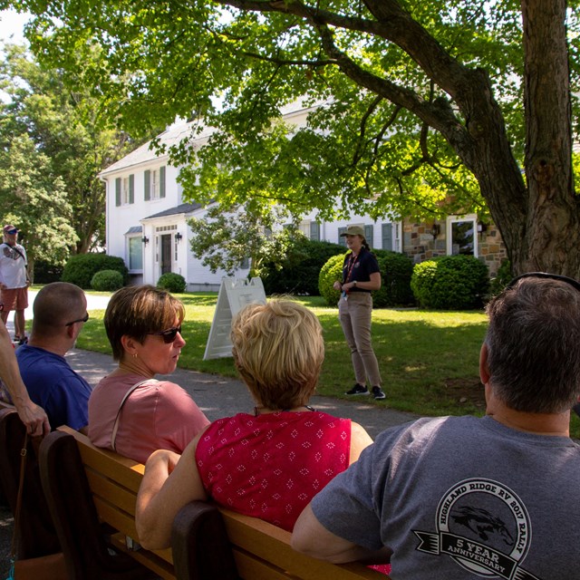 An intern giving a talk to a group of visitors in front of a large white house.