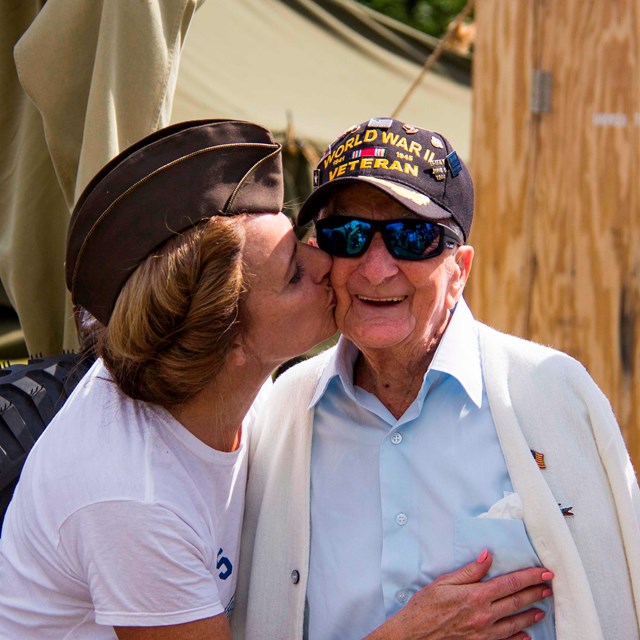 A photo of a World War II vet getting kissed by a woman on his left as another stands on the right.