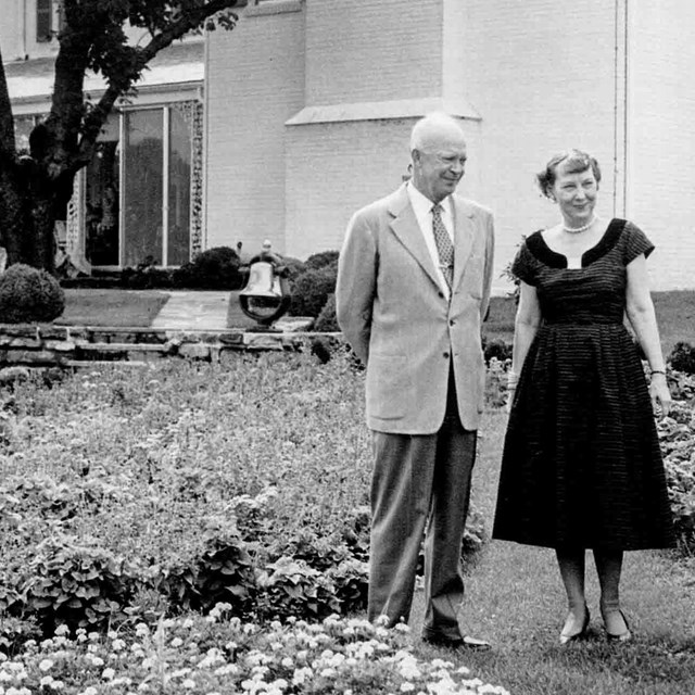 Dwight and Mamie Eisenhower walk down a path through a garden near their house. Black and white.