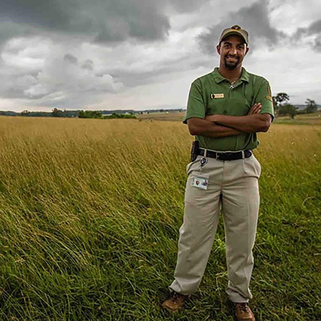 An intern poses for a picture in an open field. He's wearing a green shirt, khaki pants, & ball cap.
