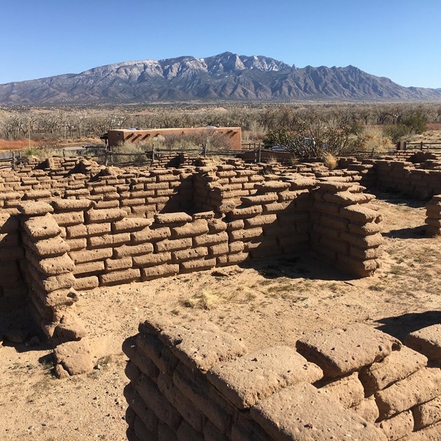 Ruins of adobe brick walls lead to a distant view of a high desert mountain capped with snow.