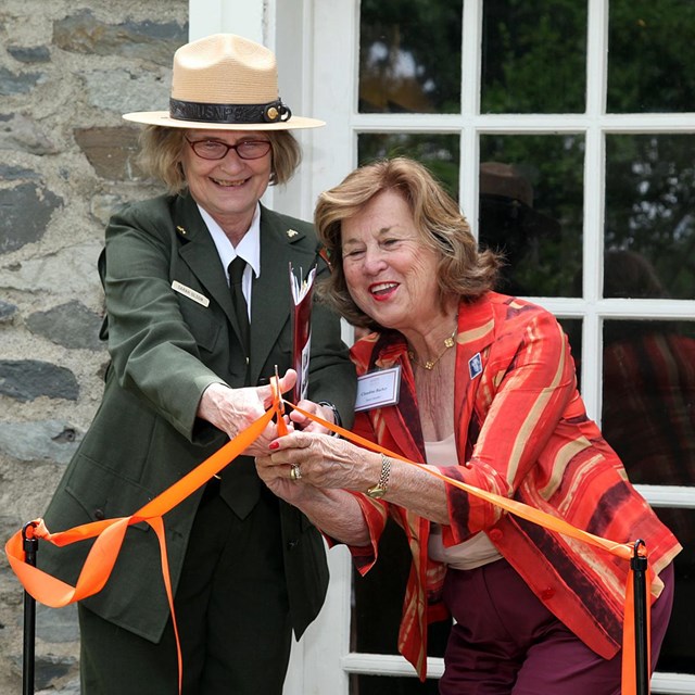 A park ranger and a woman hold a pair of scissors, cutting a ribbon.
