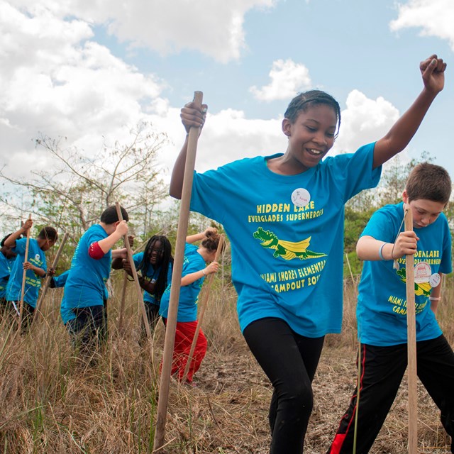 Young girl raising her arm in the air as she walks through grass, followed by other students