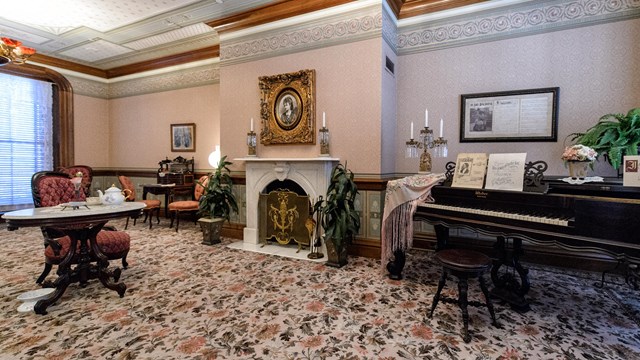 A formal parlor in a 19 century home with a piano and fireplace. 
