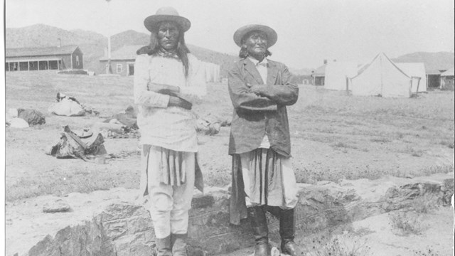 And historic black and white photo. Two Indigenous people stand on a rock wall, their arms folded.