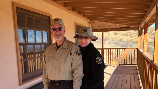 Two people in uniform stand a porch with a chair next to them.