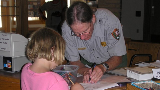 A ranger leans across a desk working on a paper with a young blonde girl in a pink shirt.