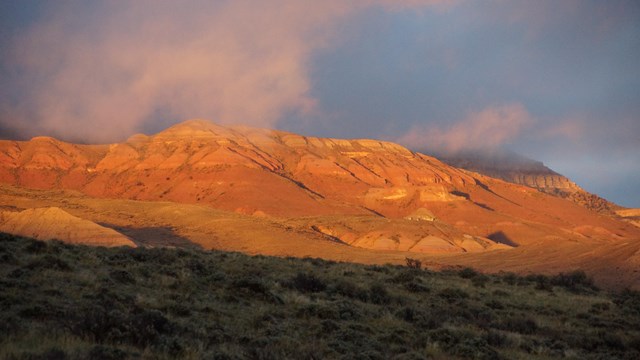 Wasatch ridgeline with red and brown is illuminated by evening light. Wisps of clouds cover the top.