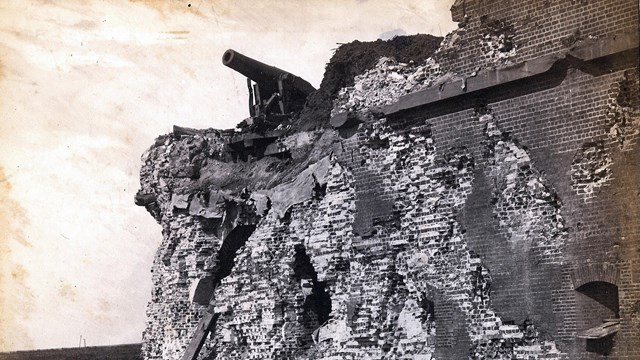 Black and white image of a damaged brick fort with a cannon on top. 