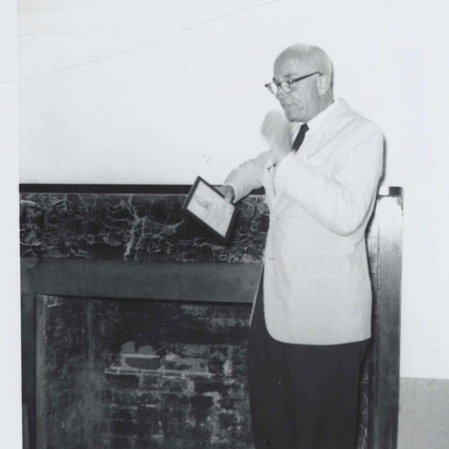 man standing next to fireplace in suit with note card