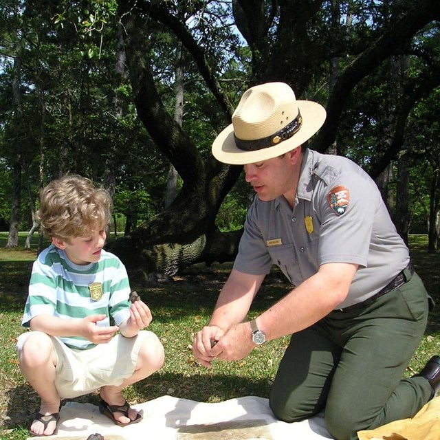 color photo of a ranger at left kneeling showing tree bark to a young boy sitting right