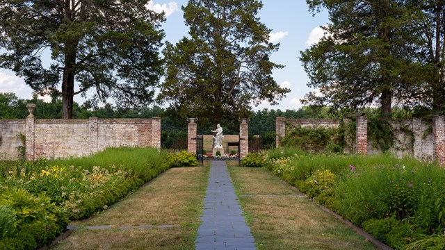 A path straight through lush, green gardens leading to a Roman statue.