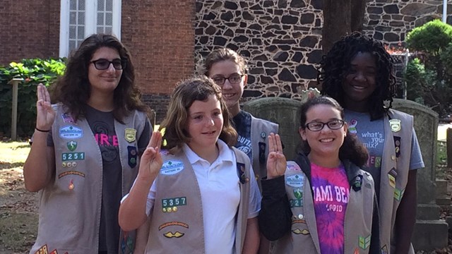 Five Girl Scouts pose in front of Old Swedes Church next to a few gravestones. 