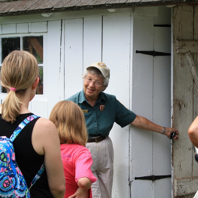 A park volunteer assists with a ranger program. She holds a barn door open for visitors to see.