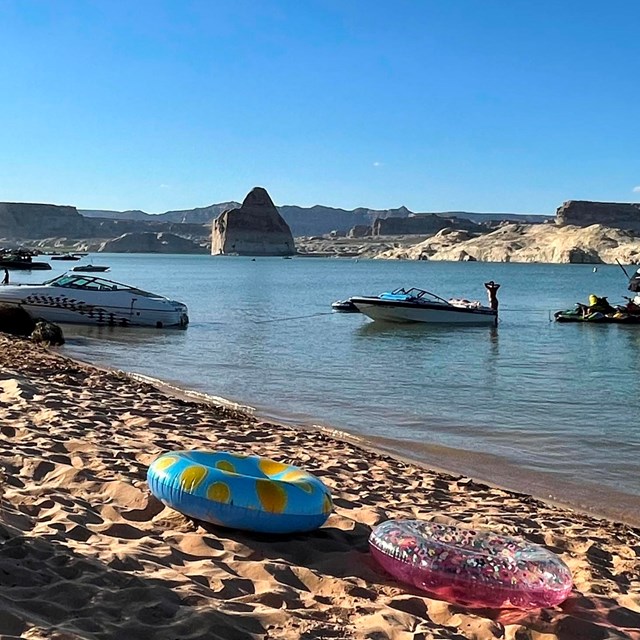 Boats and beach toys on a sandy beach with a sandstone rock in the lake behind
