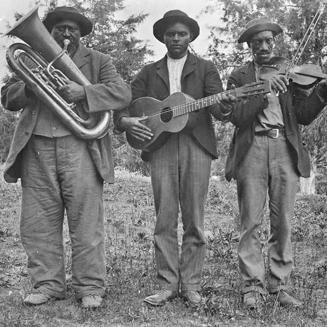 Three African American men are standing holding instruments - a tuba, guitar, and fiddle with a bow