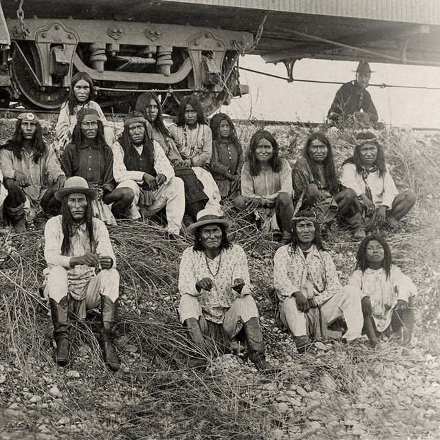Black and white photo of several American Indians sitting on a hill beside a rail car.