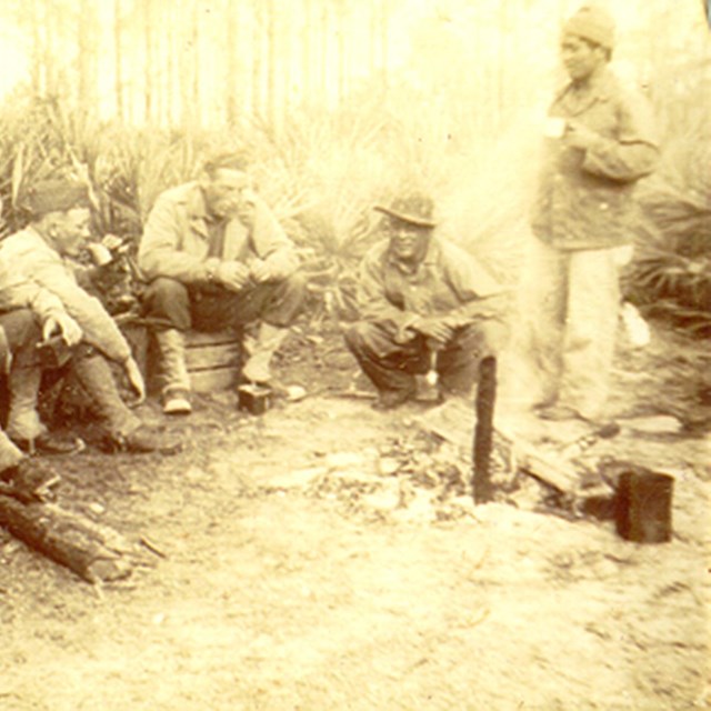 A group of soliders sit in a wooded area in a semi-circle.
