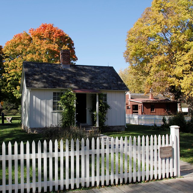A historical park features a white cottage and brown wagon shop amid colorful autumn trees.