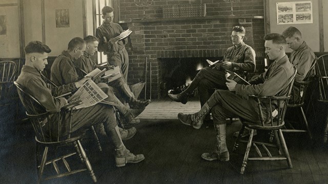 Several soldiers seated in front of a fireplace reading newspapers and books