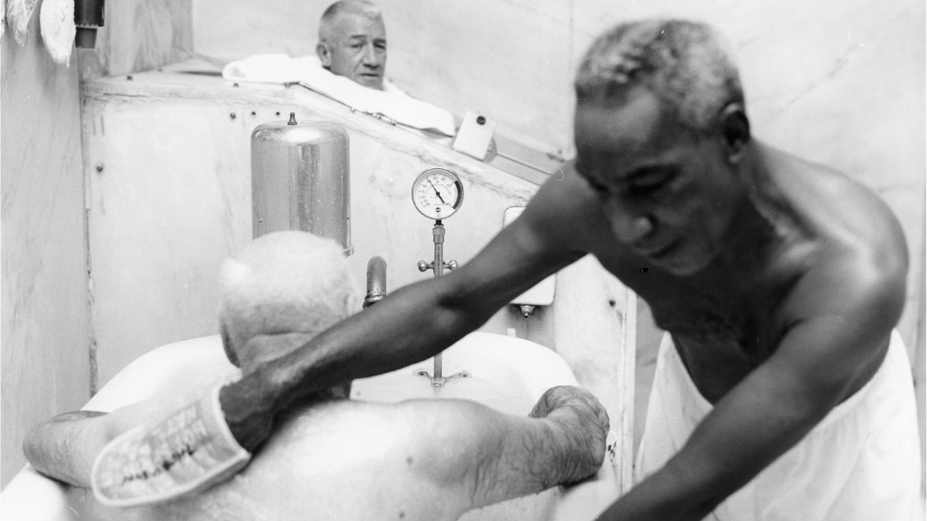 White man sits in bathtub as African American man rubs his back