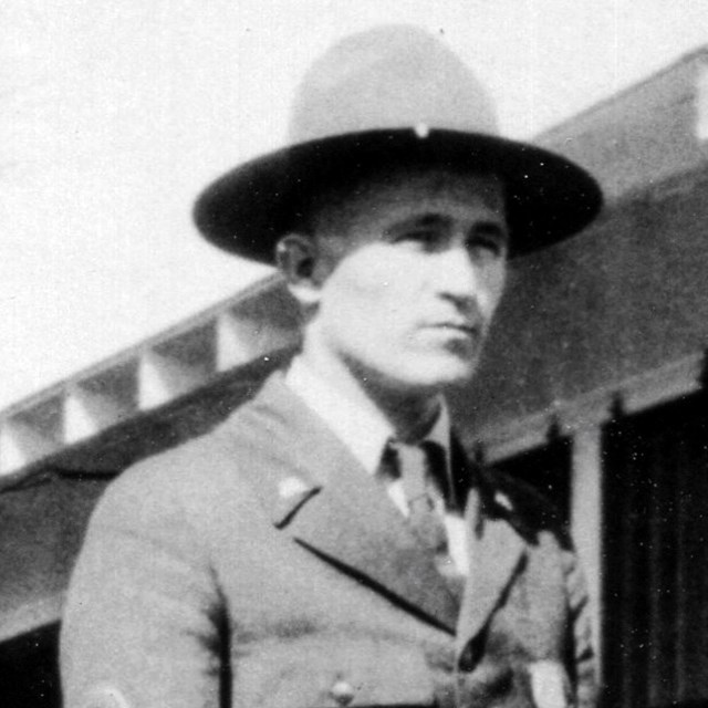 Up close image of a young man dressed in a park ranger uniform, buttoned jacket and flat hat.