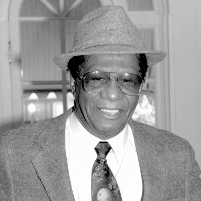 A man smiles while wearing sunglasses, a hat, a dark suit, dark tie, and white shirt.