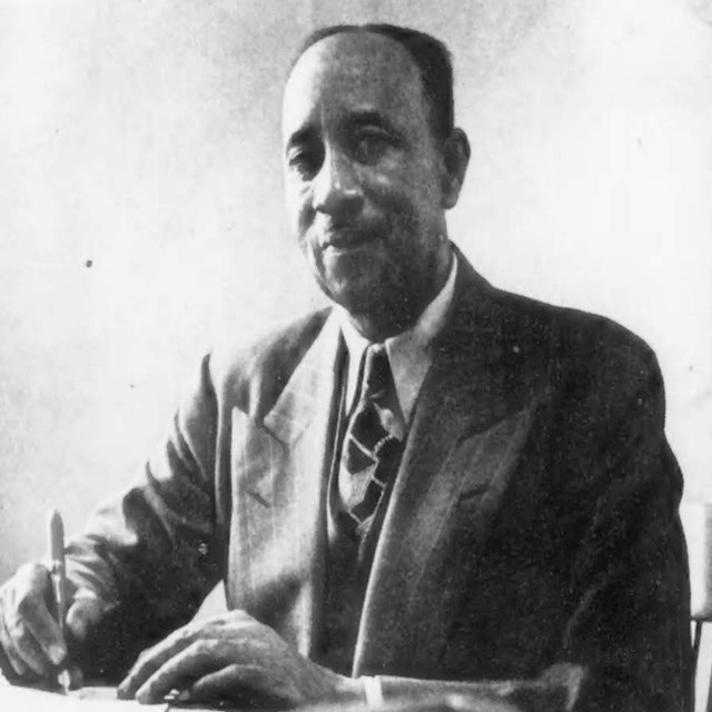 Balding man sits at table holding a pen wearing a white shirt, dark suit, and a dark tie