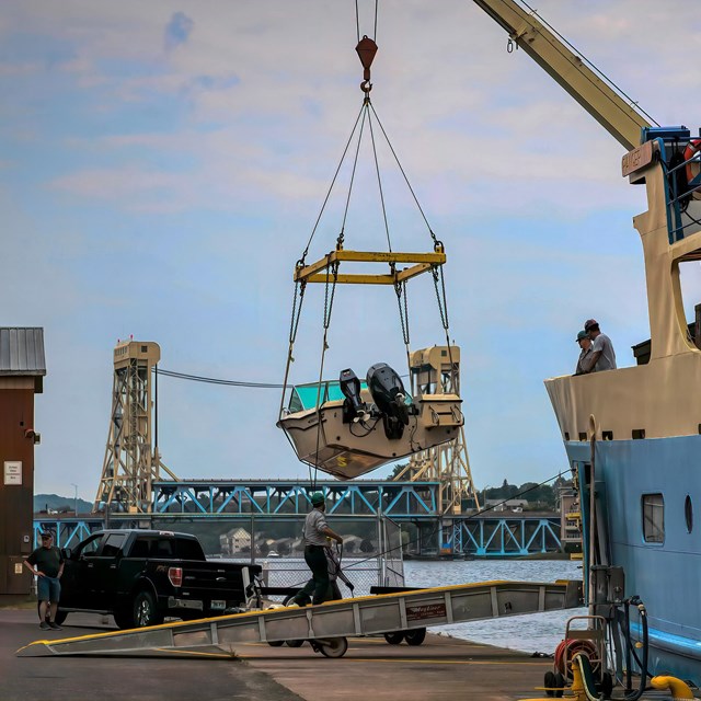 RANGER III ferry with bow crane lifting a motorboat from a truck trailer. The boat hangs in the air.