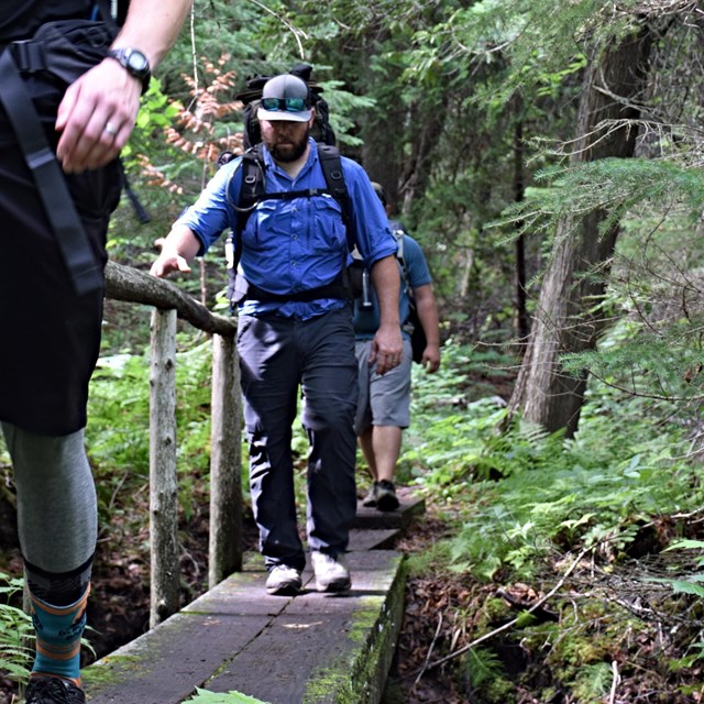 Two people with backpacks on walk across a wooden bridge in the forest.