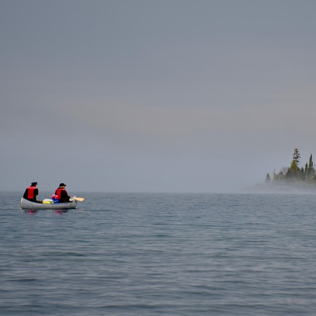 A silver canoe paddles toward an island surrounded by fog on a large body of water.