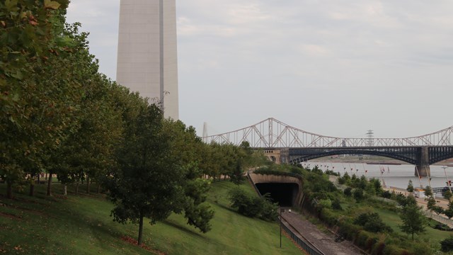 A leg of the Gateway Arch emerges from some trees to left of railroad tracks river and two bridges.