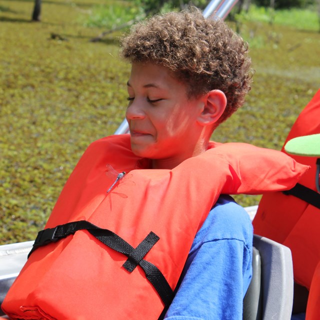 Boy with brown curly hair smiles down at a dragon fly on his chest. He wears an orange life jacket.
