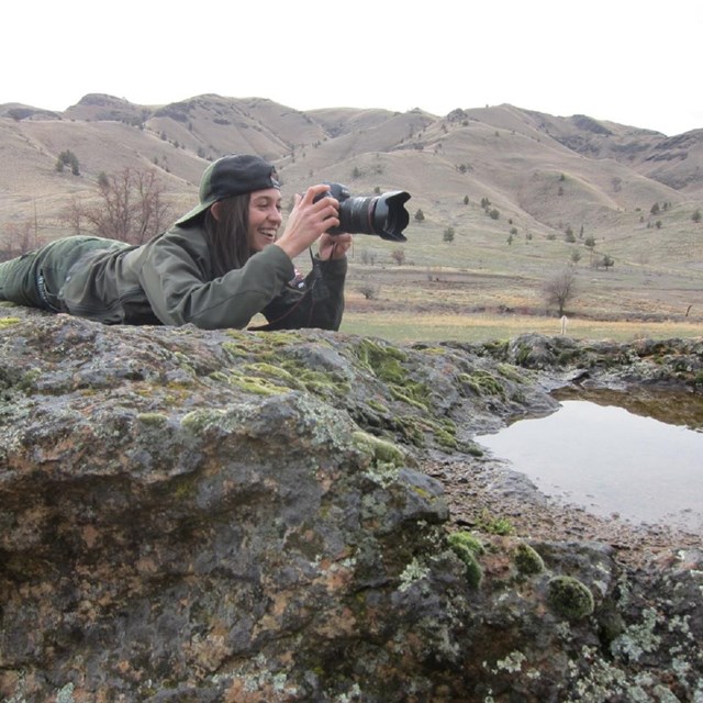 A uniformed Park Ranger smiles while taking a picture laying on top of a boulder.