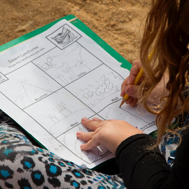 a child sitting on the ground draws desert landforms on a worksheet