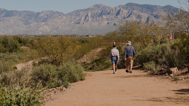 Two people walk down a dirt trail in a desert landscape toward mountains