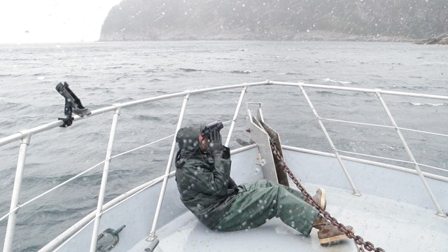 A person in full rain gear takes photos from the bow of a boat in wet conditions.