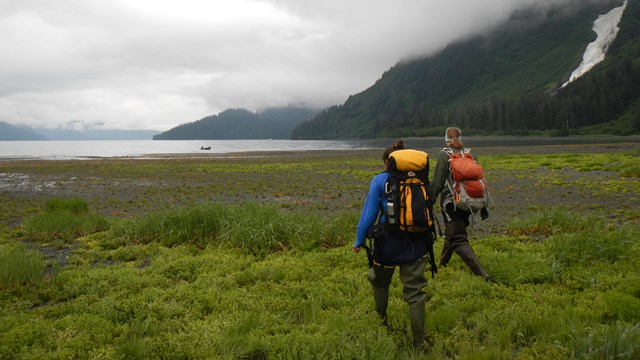 Two people with backpacks walk towards water and mountains.