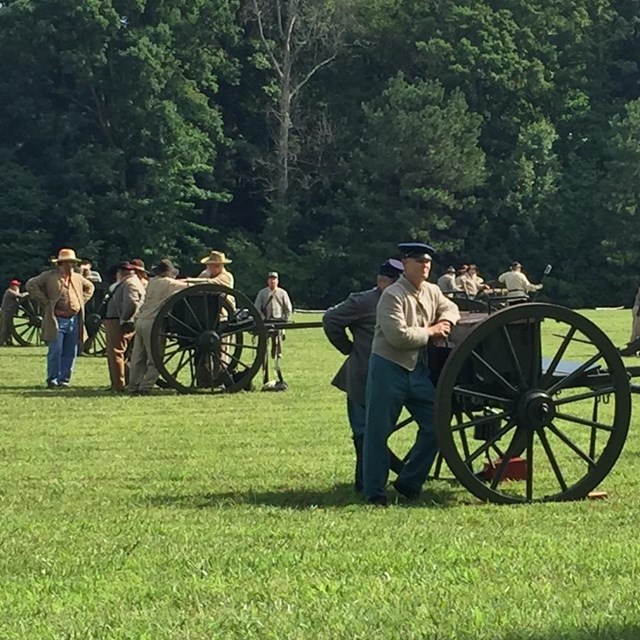 About 25 men in period costume stand in groups near 6 vintage cannons on a large grass field.