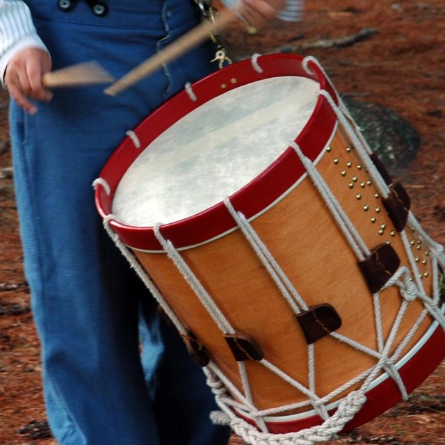 Hands use drumsticks on a large cylindrical drum while carrying it over shoulder. 
