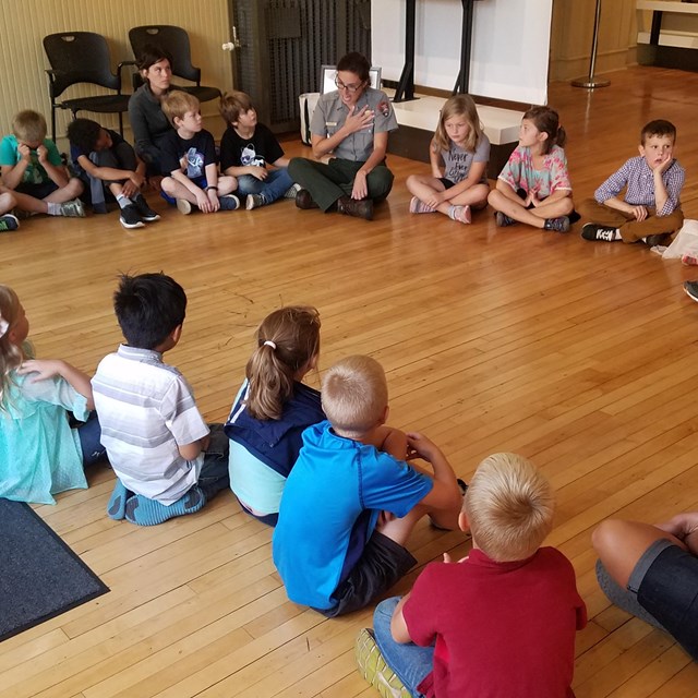 Students sit in a circle on a wood floor listening to a park ranger talk.