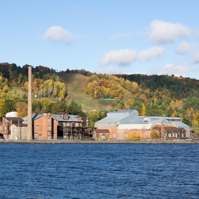 An industrial site with red brick and sandstone buildings sits on the waterfront, with fall foliage 