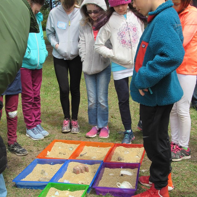 A group of students surround and look at sand in colorful trays.