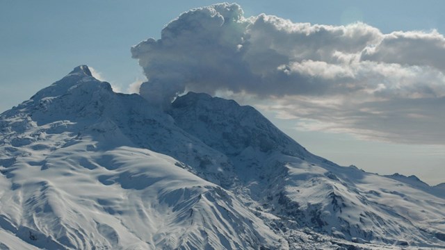 Image of a snow covered volcano steaming with clear skies in the background.