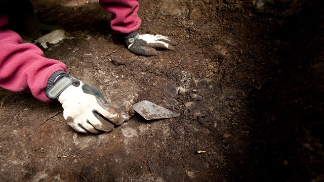 a close-up of gloved hands working digging with a trowel at an archeological site