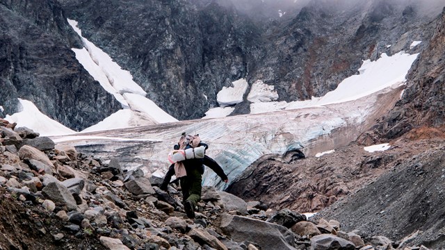 Image of a backpacker going up a talus slope towards the face of a glacier.