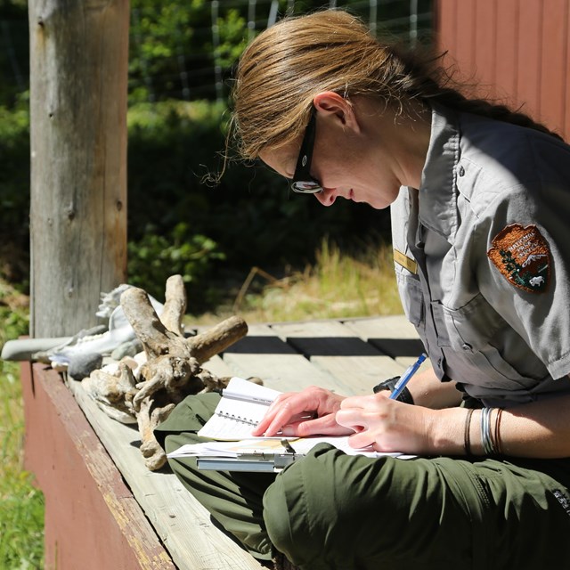 A female park ranger writes in a journal on a cabin porch