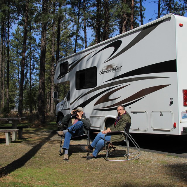 A white RV parked in a campsite with two people relaxing in camp chairs.