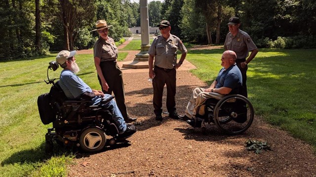 Three Park rangers talking to two visitors in wheelchairs on trail, Memorial Building in background