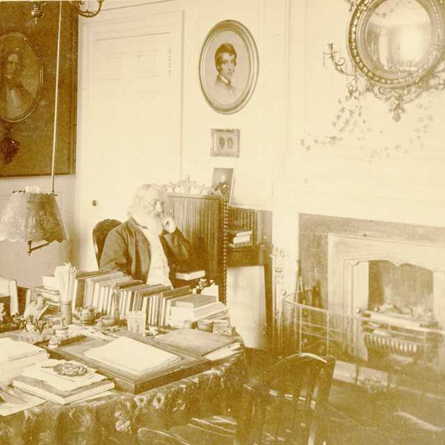 black and white photograph of man seated at table covered in books